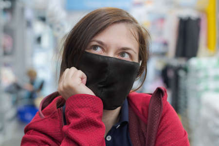 The lady in the red jacket and black protective mask was thinking about something. In the background, a shop, a clothing section can be seen in the background. Close-up shot, portrait.の写真素材