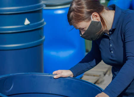 A lady in a black medical mask examines a large blue bucket. A clear sunny day, outdoors.の写真素材