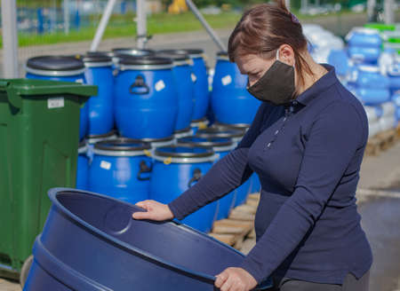 A woman in a black, medical mask examines the blue barrel. In the background pallets with plastic...
