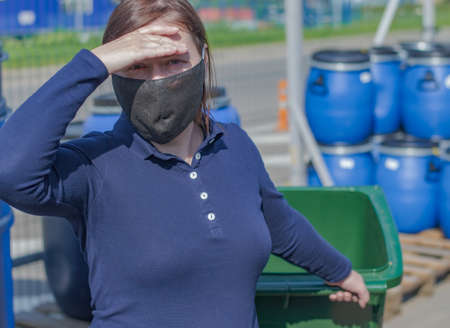 Girl in a protective mask shuts out the sun and looks at the camera. In the girls hand is a green plastic container. An iron fence is visible in the background.の写真素材