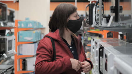 Caucasian women wearing antivirus medical mask and red jacket picks out microwave in electronics...