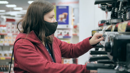 European a women wearing antivirus medical mask and red jacket picks out coffee maker in device...