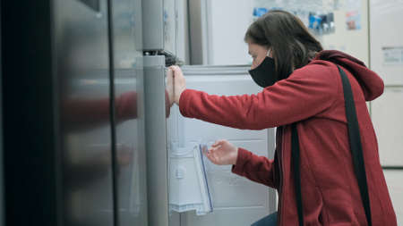A young woman wearing a black mask antivirus medical and red jacket picks out a fridge at a home appliance storeの写真素材