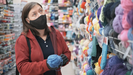 Woman wearing a mask and gloves against the virus chooses chenille yarn for knitting and needlework. The concept of hobby and craftの写真素材