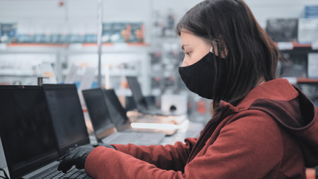 Caucasian woman wearing a mask and gloves against virus in electronics store. She stops at the laptop rack and selects a deviceの写真素材
