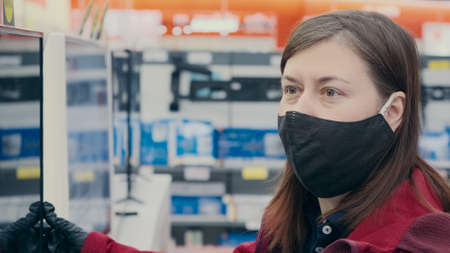 Woman in mask against virus is in an electronics store surrounded of working monitors. The concept of buying new gadgets and devicesの写真素材