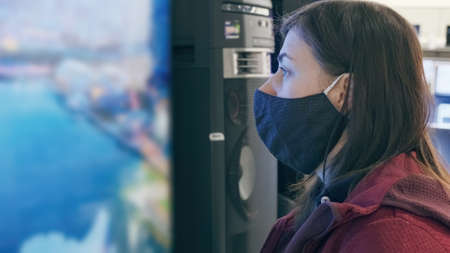 Woman wearing an against virus mask looks at a huge plasma TV in gadget and device store, next to...
