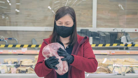 Girl wearing mask and gloves against virus buys pork meat in grocery store. She carefully examines...