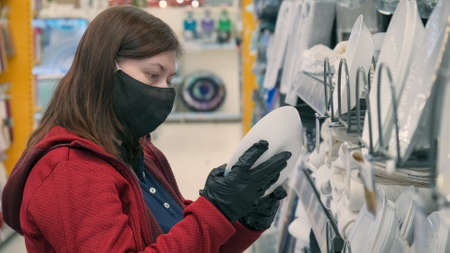 Girl in the mask and gloves against the virus buys a deep, white for food plate in a store. Rows of dishes can be seen on the shelves. Concept of safe shopping during epidemics and pandemicsの写真素材