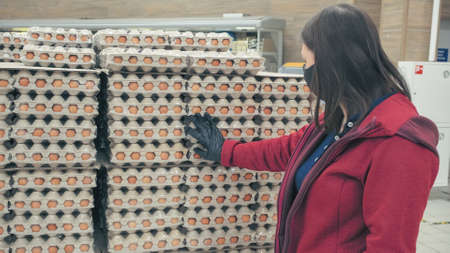 Girl wearing mask and gloves against virus buys chicken eggs in grocery store. Concept of...