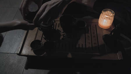 Tea master sits near board on which there are cups with drink candle and statue of Buddha. Woman...