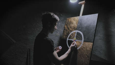 Young Caucasian man paints beautiful abstract picture in semi darkness. Easel is illuminated by one...