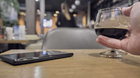 In a restaurant, there is a dark drink in a wine glass on the table, and a phone is lying next to it. Man hand picks up the glassの写真素材