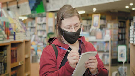 Serious woman wearing glasses and mask against virus stands in second hand bookstore and makes notes...