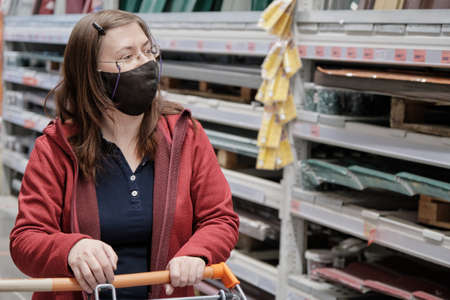 Caucasian woman with glasses rides through building materials store with grocery cart. She wears mask against virus. Concept of women independence, gender equality, repair and constructionの写真素材