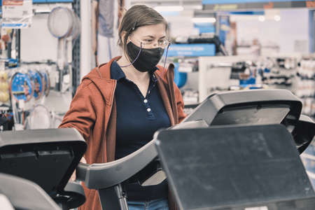 Serious woman in glasses and a mask against the virus buys a large treadmill in a store. The concept of sports and a healthy lifestyleの写真素材