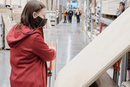 Woman in mask against virus is in building materials store she holds cart on which huge sheet of...
