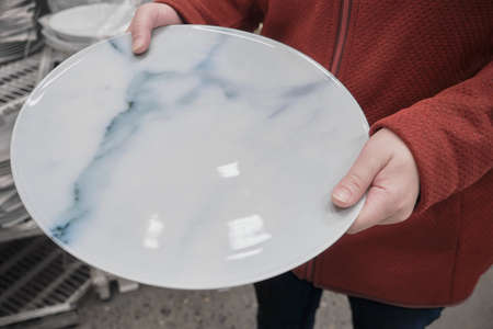 Caucasian woman buys a large white plate with a marble pattern in a store for serving the table. The concept of buying new dishes and kitchen equipment. Hands close up shotの写真素材