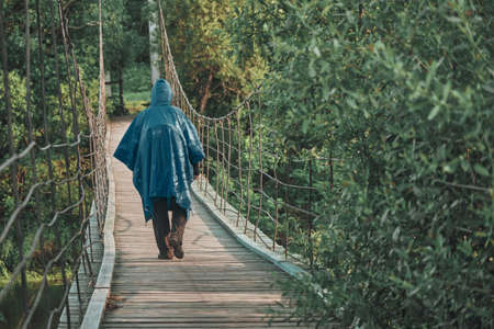 Girl with backpack under blue raincoat is engaged in hiking. On summer evening at sunset, she walks along wooden suspension bridge over river. Concept of savage travel, lifestyle, outdoor recreationの写真素材