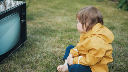 Small child in yellow jacket is sitting on grass and watching an old retro TV. TV is strobing. Concept of post apocalypse, horror, surrealism and mysticism. Children dependence on TV, gadgets devicesの写真素材