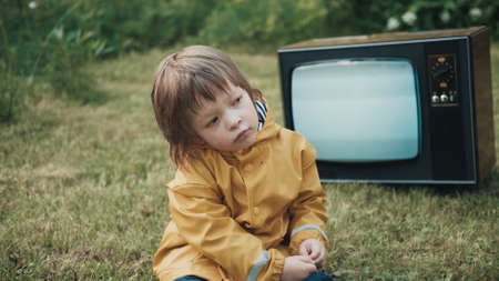 Small, sad child dressed in a yellow raincoat is sitting on the grass. Behind him, an old, retro TV is working. Concept of post apocalypse, horror, surrealism and mysticismの写真素材