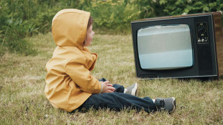 Small, cute child, dressed in a yellow raincoat with a hood, is watching an old, vintage, retro TV and smiling. There is interference on TV in the form of a strobe. Concept of post apocalypse, horrorの写真素材