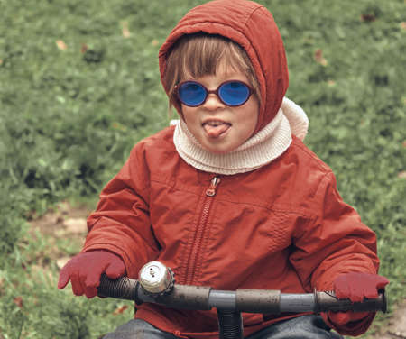 Little girl in a hood and blue glasses on a small bike. The child shows his tongue. The concept of childhood, humor and lifestyle. Close up portrait in retro styleの写真素材