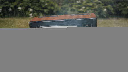 Old vintage retro TV stands on the grass. Smoke billows from the damaged device. Concept of post apocalypse, surrealism and the fire hazard of old worn out equipmentの写真素材