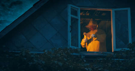 Maniac in a yellow raincoat with an axe and a knife on the top floor of an old house. The concept of horror, thriller and Halloween holiday. At night, the camera shoots outside through the windowの写真素材