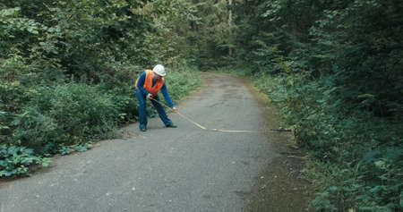 Worker in workwear and a helmet on the highway in the forest measures the roadway with a tape measure. The concept of repairing roads, a fallen tree and an accident on a forest, country roadの写真素材