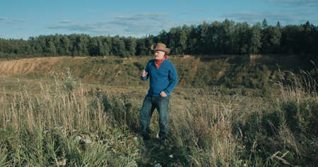 Man in a cowboy hat stands on the edge of a sandy ravine overgrown with forest and smokes a cigarette. There is wild nature around, it is eveningの写真素材