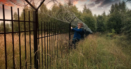 Man runs to the fence with barbed wire and climbs on it. Evening sunset. The concept of illegal border crossing, penetration into a protected object and unlawful migrationの写真素材