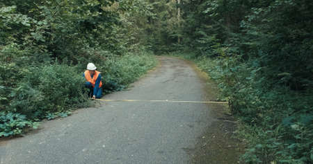Worker in workwear and a helmet on the highway in the forest measures the roadway with a tape measure. The concept of repairing roads, a fallen tree and an accident on a forest, country roadの写真素材