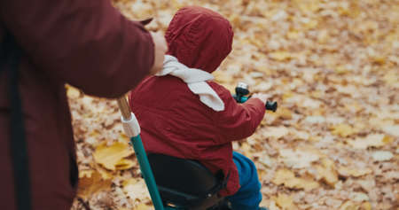 Mom rides cute little girl on bicycle in autumn park. Concept of childhood, happy family and lifestyle. Nature, clean air, outdoor activities on weekends. Cool weather, around leave of Canadian mapleの写真素材