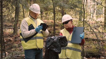 Man and woman environmentalists in workwear and helmets document offense - forest pollution and issue fine. Concept of pollution of natural parks, woods and nature reserves with non decomposable wasteの写真素材