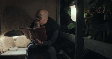 An angry elderly man is sitting on porch at night, he is reading a book using magnifier glass. Concept of a scary fairy tale, horror, nightmares and black humor. Old, retro decor, burlap backgroundの写真素材