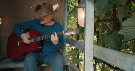 Man cowboy with acoustic guitar is sitting on porch of farmhouse, playing an instrument and singing songs. Concept of creativity, hobbies, lifestyle and relaxation. Old, retro decor, burlap backgroundの写真素材