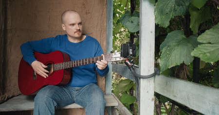 Man with an acoustic guitar sits on porch of village house and hosts an online podcast using his phone. Concept of creativity, hobbies, lifestyle and recreation. Old, retro decor, burlap backgroundの写真素材