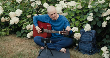 Man with an acoustic guitar is sitting in a park in nature and tuning the instrument using an app on his phone. The concept of travel, hiking and hobbiesの写真素材