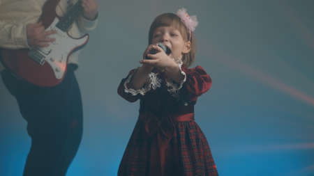 Little girl on stage in vintage dress, she sings into microphone and dances, her father plays an electric guitar. Color music is shining and smoke is billowing. Family time, creativity and hobbiesの写真素材