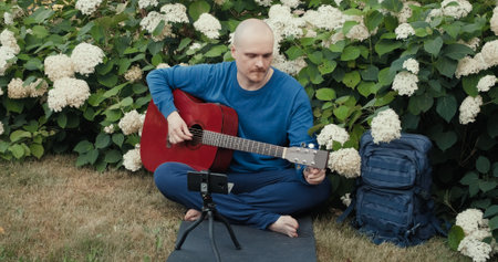 Man with an acoustic guitar is sitting in a park in nature and tuning the instrument using an app on his phone. The concept of travel, hiking and hobbiesの写真素材