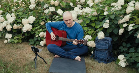 Man in with an acoustic guitar is sitting in a park in nature and is streaming online podcast using gadgets. The concept of travel, hiking, internet streams, hobbies and remote workの写真素材