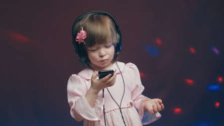 Cute little girl in big headphones and a beautiful pink vintage dress is listening to music on her phone. There is a color music in the background. The concept of music mania and audiophiliaの写真素材