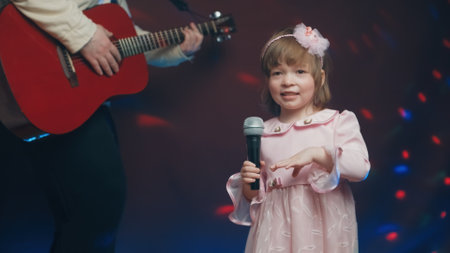 Little girl on stage in vintage dress, she sings into microphone and dances, her father plays acoustic guitar. Color music is shining and smoke is billowing. Father and Child Day, performance on stageの写真素材