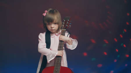 Cute little girl stands on stage in vintage dress and plays an acoustic guitar like a counter bass or cello. The concept of performing the author jazz and blues music. Hobbies and art among childrenの写真素材