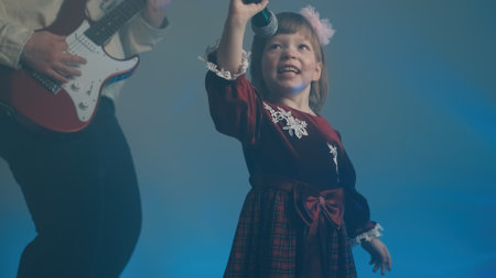 Little girl on stage in vintage dress, she sings into microphone and dances, her father plays an electric guitar. Color music is shining and smoke is billowing. Family time, creativity and hobbiesの写真素材