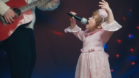 Little girl on stage in vintage dress, she sings into microphone and dances, her father plays acoustic guitar. Color music is shining and smoke is billowing. Father and Child Day, performance on stageの写真素材