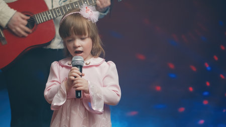 Little girl on stage in vintage dress, she sings into microphone and dances, her father plays acoustic guitar. Color music is shining and smoke is billowing. Father and Child Day, performance on stageの写真素材