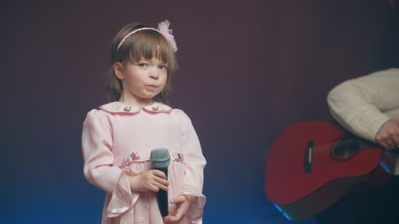 Little girl on stage in pink, vintage dress. In background, father with acoustic guitar adjusts musical equipment on stage. Color music is shining. Performance on stage. Early development of childrenの写真素材