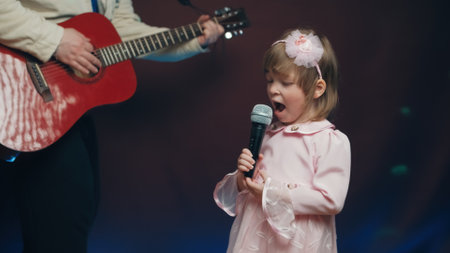 Little girl on stage in vintage dress, she sings into microphone and dances, her father plays acoustic guitar. Color music is shining and smoke is billowing. Father and Child Day, performance on stageの写真素材